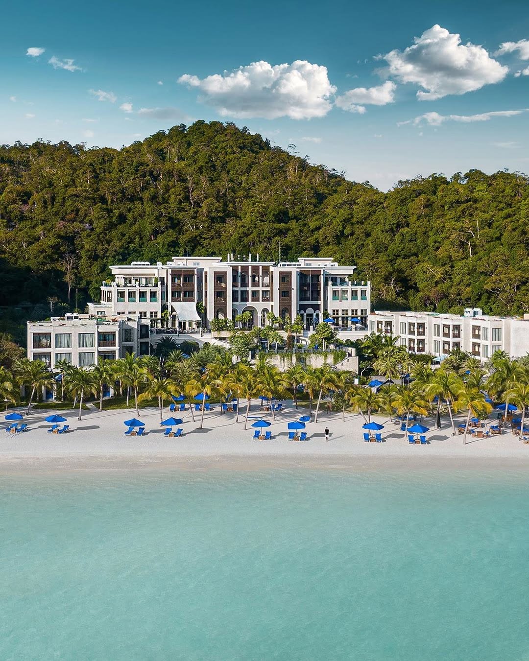Aerial view of The St. Regis Langkawi beachfront resort with grand architecture, palm-lined beach, blue umbrellas, and lush rainforest backdrop.