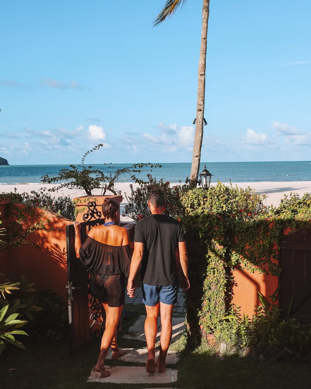 Couple walking through a garden gate at Casa del Mar Langkawi towards a sandy beach and calm sea.