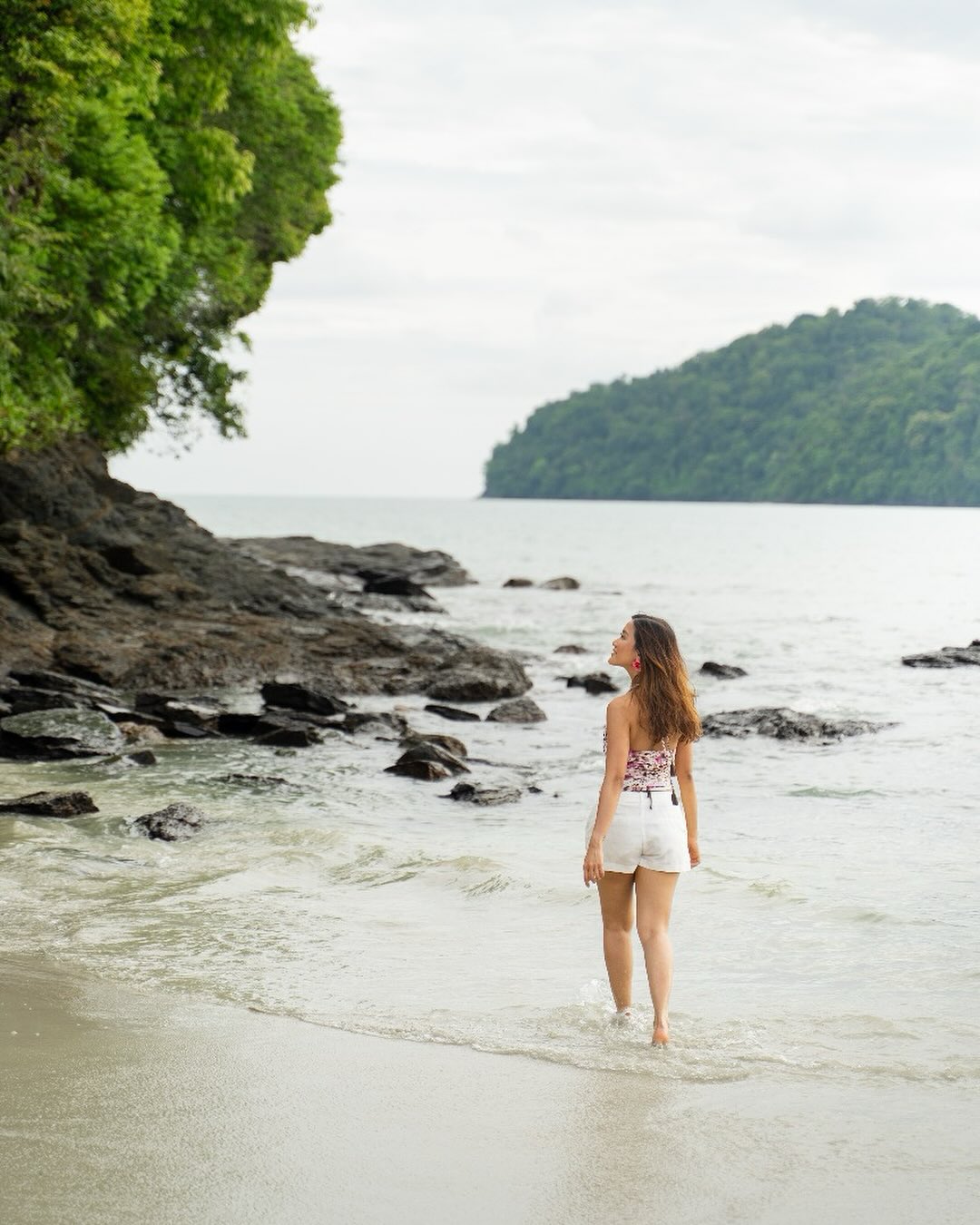 Woman walking along the shoreline at Aloft Langkawi Pantai Tengah with rocky coast, calm sea, and lush greenery.