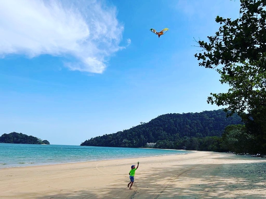 Child flying a kite on a quiet beach at The Datai Langkawi with clear blue sea, sandy shore, and forested hills in the background.