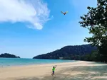 Child flying a kite on a quiet beach at The Datai Langkawi with clear blue sea, sandy shore, and forested hills in the background.