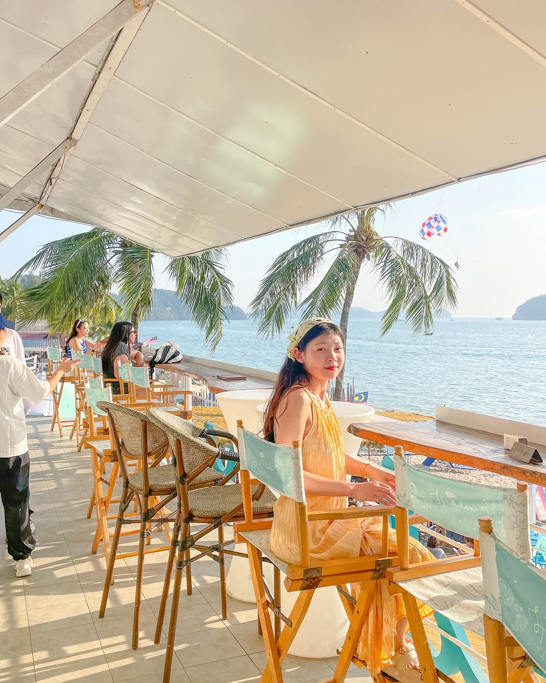 People seated at a beachfront café at Pantai Tengah Langkawi enjoying sea views with palm trees and parasailing in the distance.