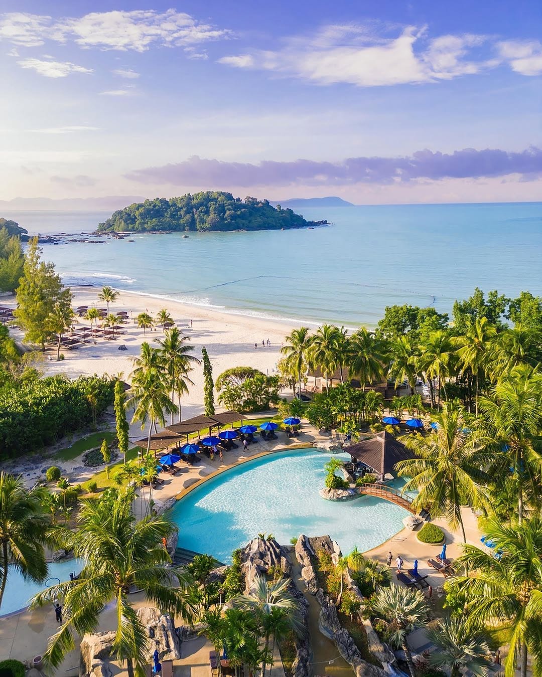 Langkawi resort pool surrounded by palm trees overlooking a sandy beach and a small island in the sea.