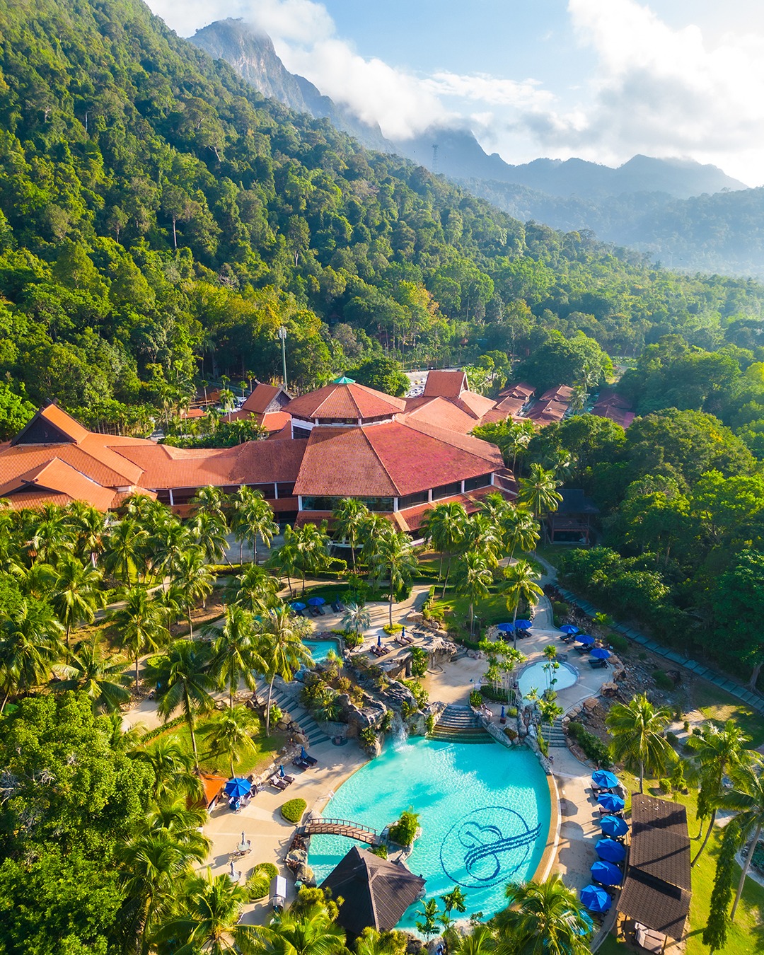 Berjaya Langkawi Resort featuring a tropical beachfront resort surrounded by lush rainforest-covered mountains, expansive lagoon-style swimming pools, swaying palm trees, and traditional Malay-inspired architecture.
