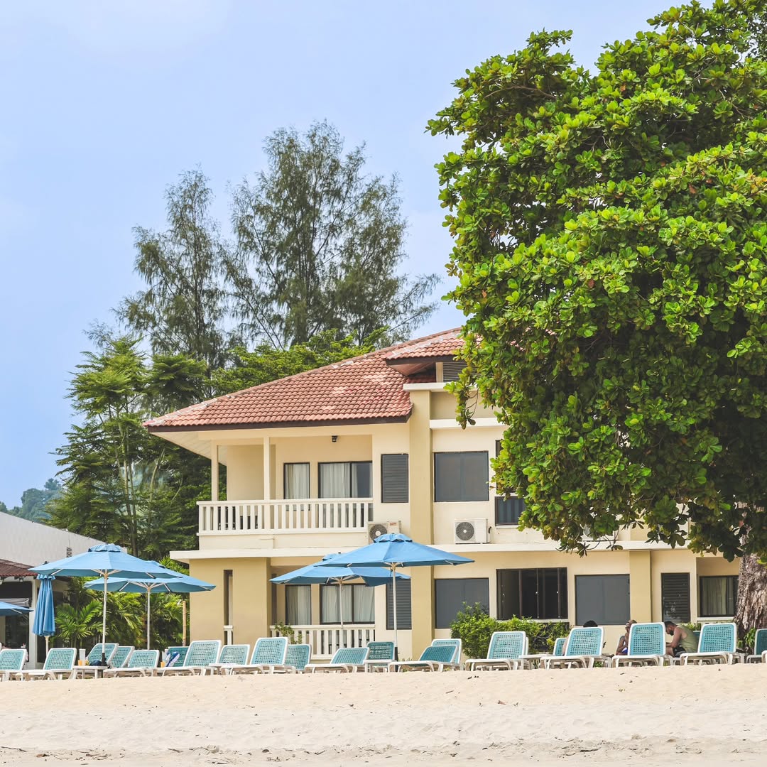 Camar Resort Langkawi featuring a beachfront resort with low-rise tropical accommodations, direct beach access, blue sun loungers and umbrellas, mature shade trees, and a relaxing seaside atmosphere along Pantai Tengah.