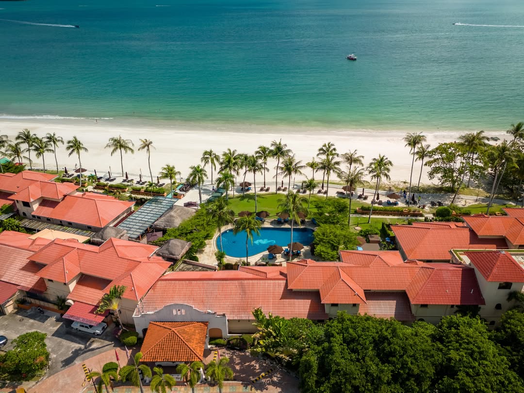 Casa del Mar Langkawi featuring an aerial view of a beachfront resort with red-tiled roofs, tropical palm trees, a freeform swimming pool, a white sandy beach, and turquoise waters along Pantai Cenang.