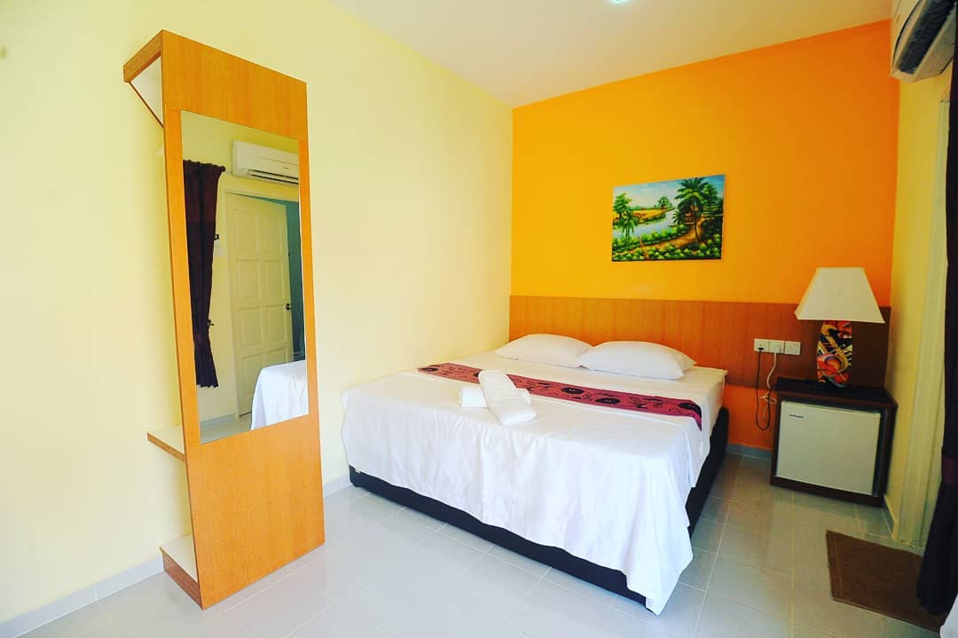 Chenang Inn Langkawi featuring a cozy guest room with a king-size bed, warm orange accent wall, simple wooden furnishings, tiled flooring, and a bright, comfortable atmosphere.