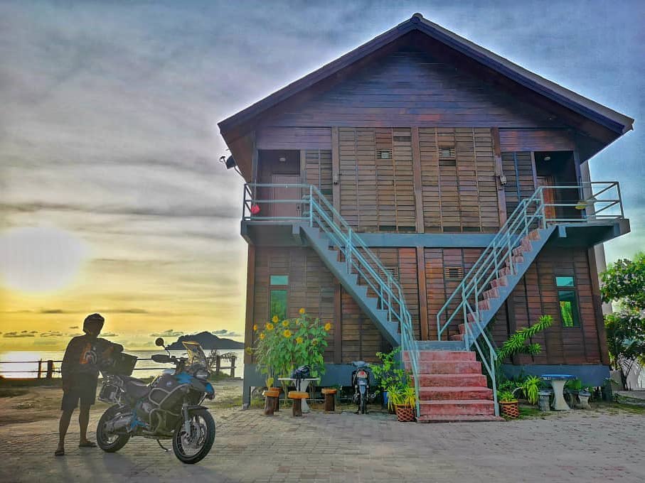 De Balqis Chalet Tanjung Rhu Langkawi featuring a traditional two-story wooden chalet with twin exterior staircases, set near the beachfront at sunset with a motorcycle parked in front and a peaceful coastal atmosphere.