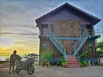 De Balqis Chalet Tanjung Rhu Langkawi featuring a traditional two-story wooden chalet with twin exterior staircases, set near the beachfront at sunset with a motorcycle parked in front and a peaceful coastal atmosphere.