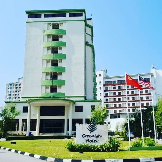 De Greenish Village Langkawi featuring a modern mid-rise hotel with green accents, landscaped gardens, national flags, and a convenient location in Kuah Town close to shopping and local attractions.
