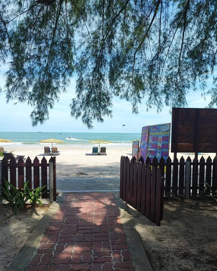 Delta Chenang Resort Langkawi featuring a shaded garden pathway leading directly to Pantai Cenang Beach, with lounge chairs, golden sand, turquoise waters, and a tranquil tropical seaside setting.
