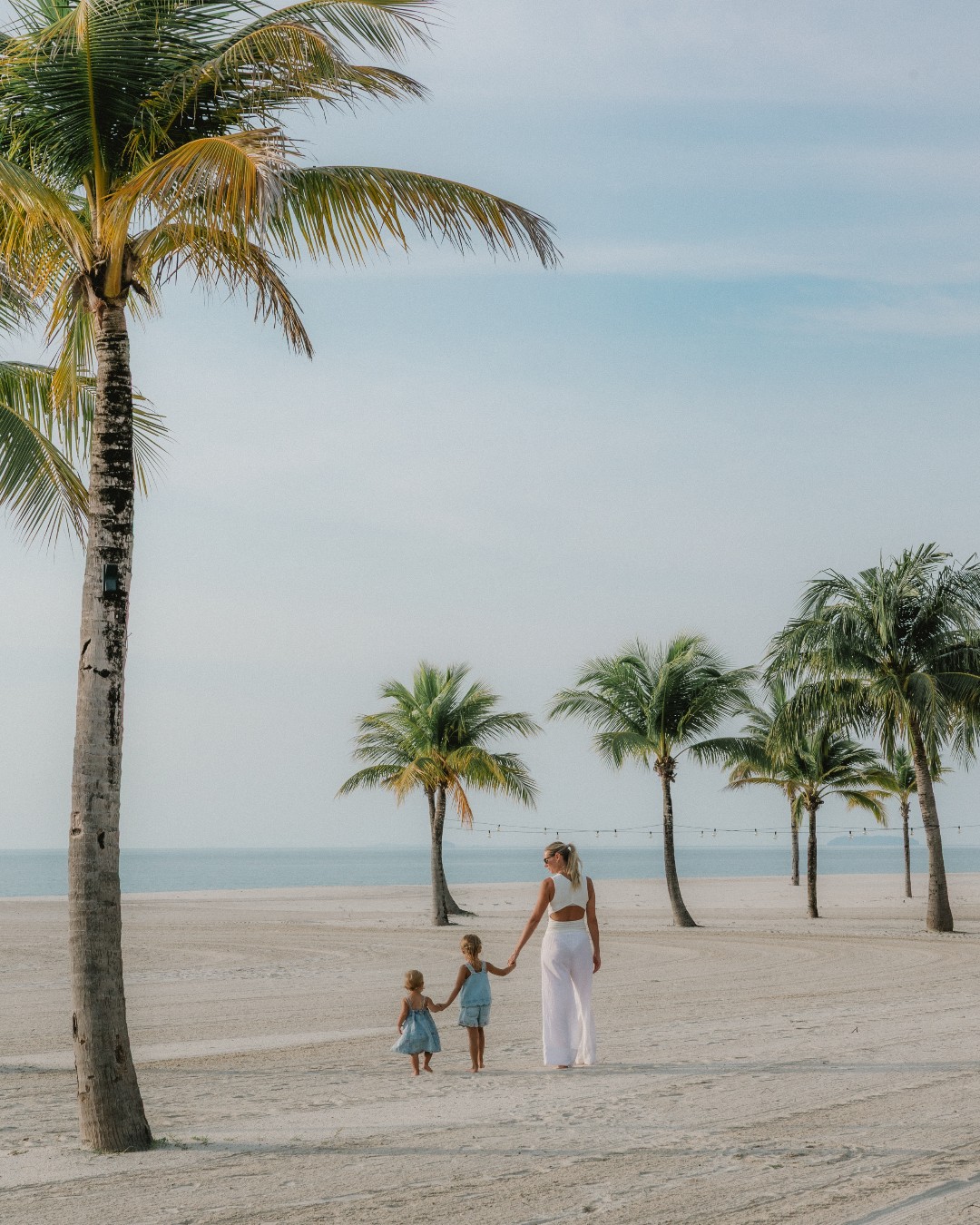 A family walking along a sandy beach at Four Seasons Resort Langkawi with palm trees and calm sea in the background.