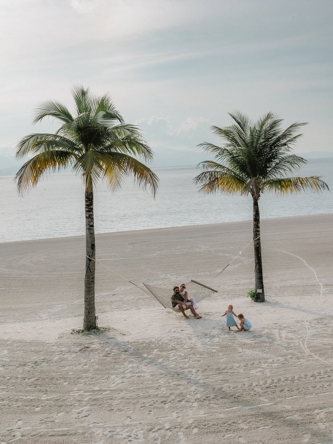 Family relaxing on a hammock between palm trees at Four Seasons Langkawi with children playing on the sandy beach by the sea.