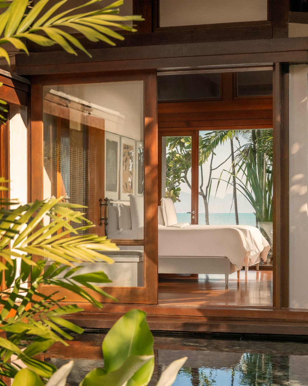Four Seasons Resort Langkawi featuring a luxurious beachfront villa with warm timber architecture, floor-to-ceiling glass doors, elegant bedroom interiors, lush tropical landscaping, and serene ocean views.
