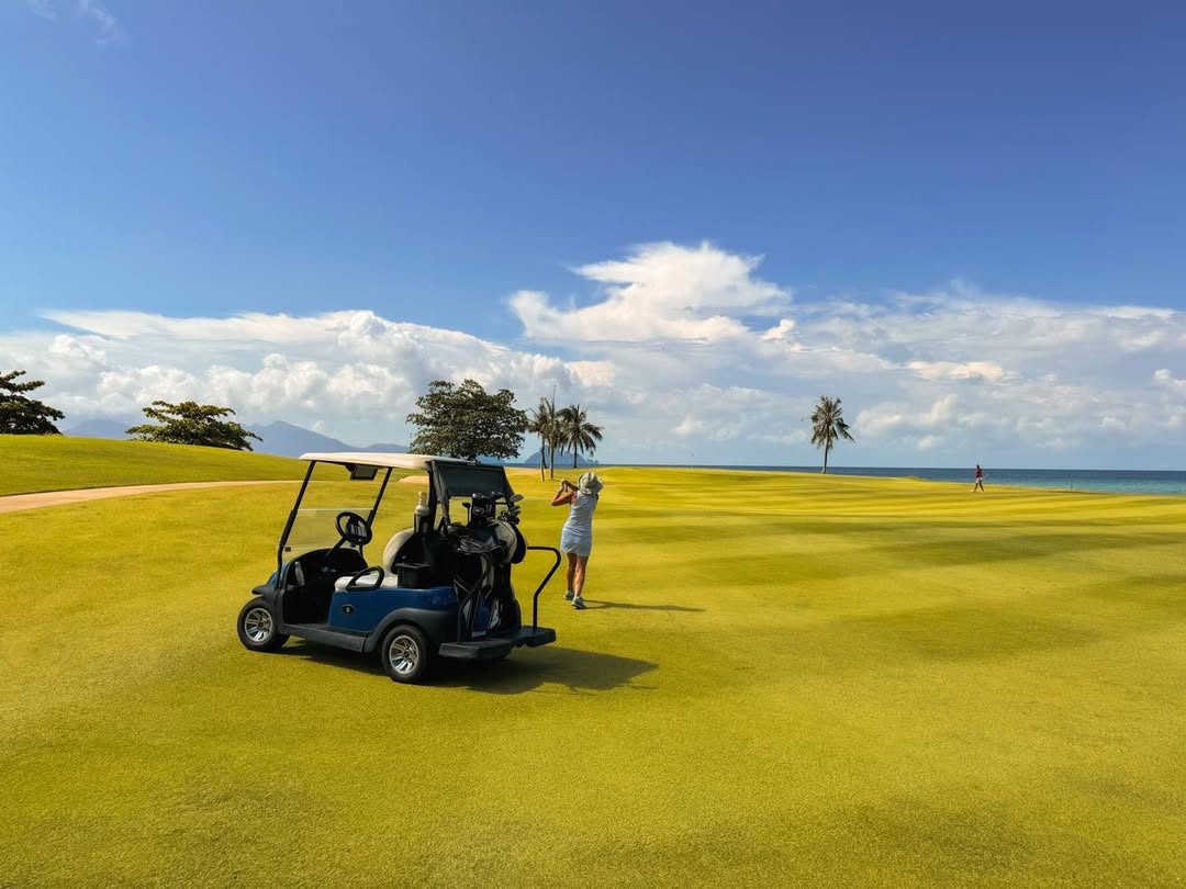 Golfer teeing off at Els Club Teluk Datai Langkawi with ocean views, manicured greens, and a golf cart nearby.