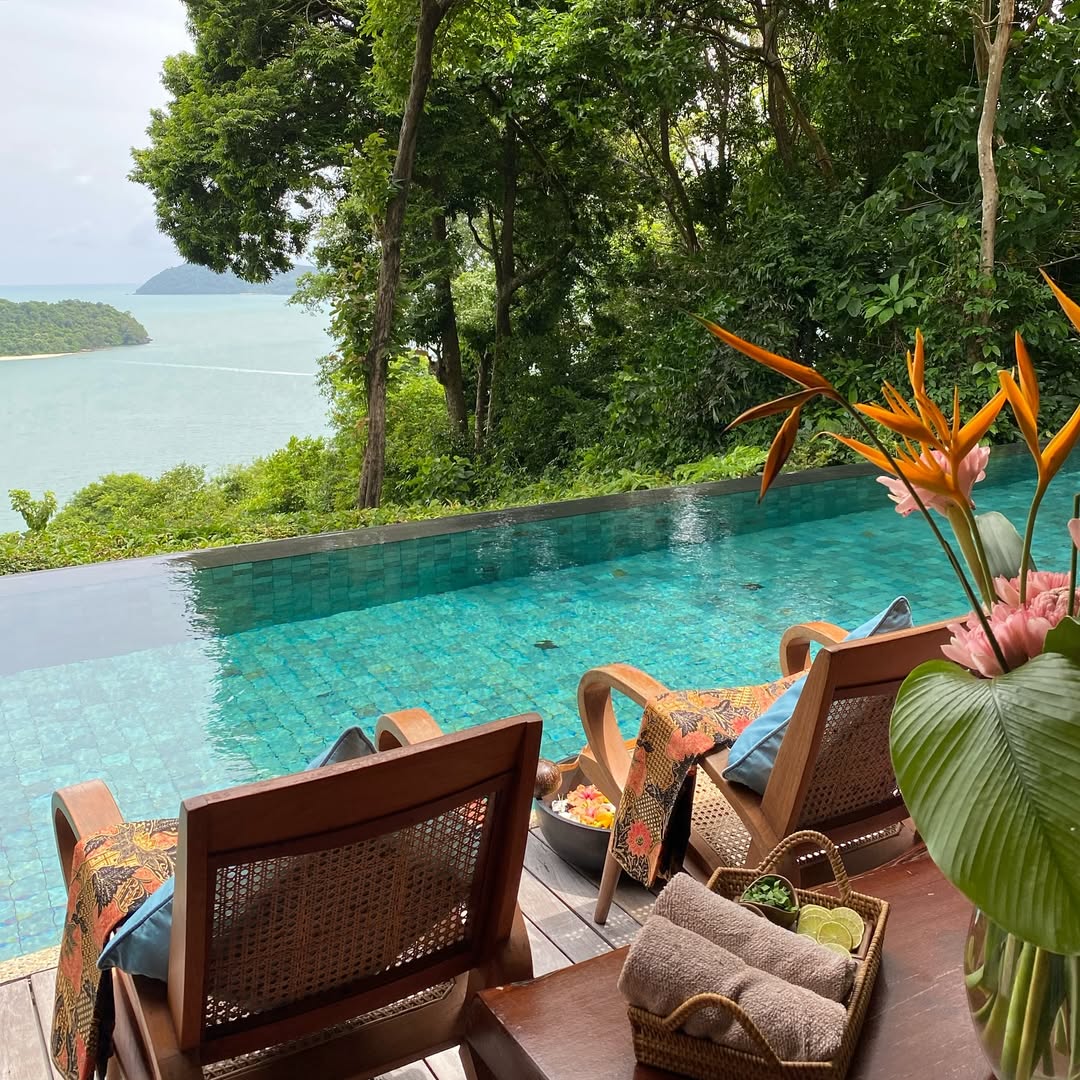 Infinity pool at Ambong Pool Villas Langkawi overlooking lush rainforest and the sea, with lounge chairs and tropical decor.