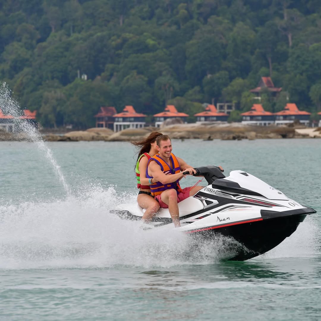 Two people riding a jet ski on the sea in Langkawi with resort villas and lush hills in the background.