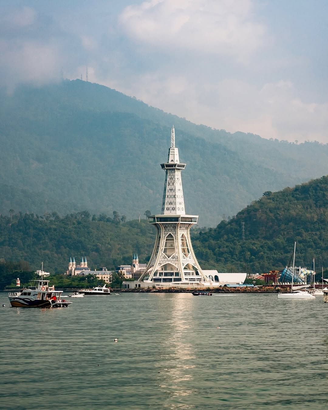 MAHA Tower in Langkawi standing by the waterfront with boats on the sea and forested hills in the background.