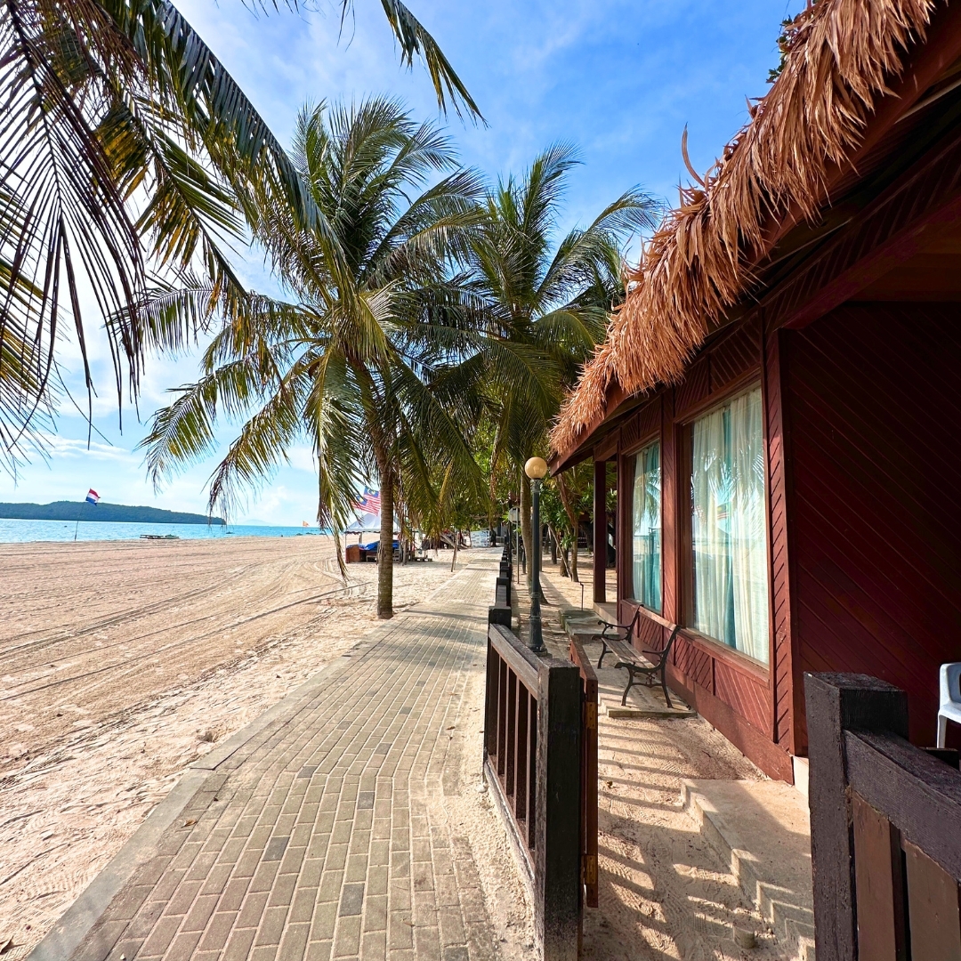 Malibest Resort Langkawi featuring traditional wooden beachfront chalets with thatched roofs, swaying coconut palms, a sandy beach, and a scenic seaside boardwalk along Pantai Cenang.