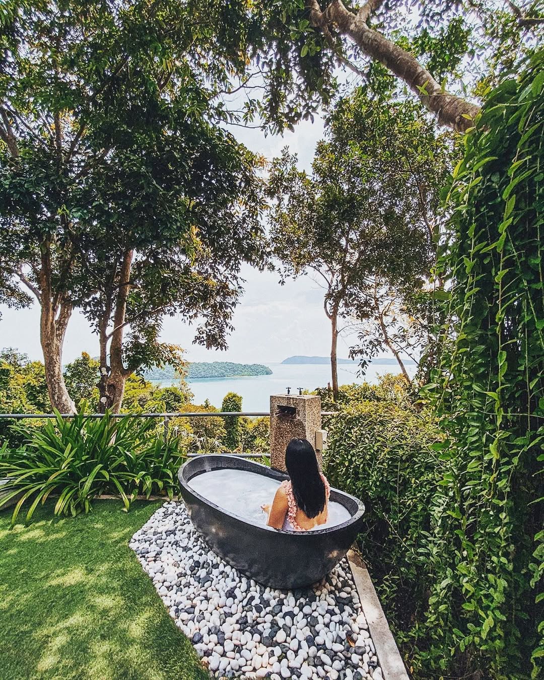 Person relaxing in an outdoor bathtub at Ambong Pool Villas Langkawi surrounded by lush greenery with a view of the sea and nearby islands.