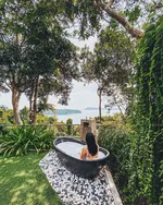 Person relaxing in an outdoor bathtub at Ambong Pool Villas Langkawi surrounded by lush greenery with a view of the sea and nearby islands.