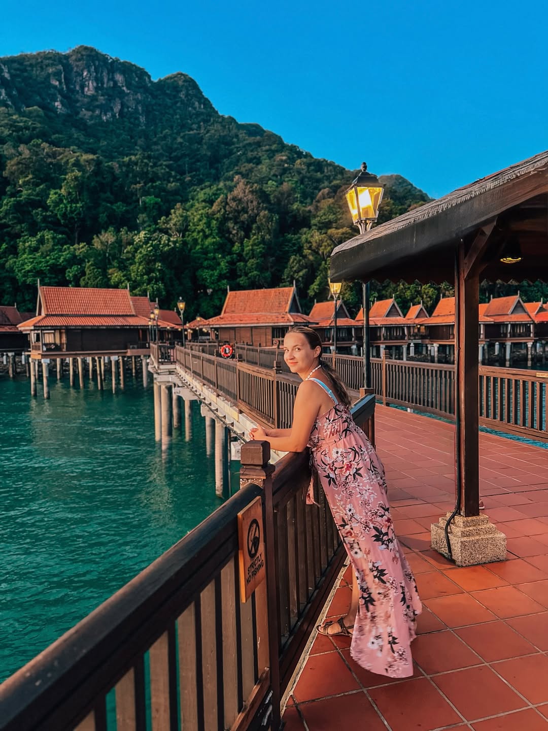 Woman standing on a boardwalk at Berjaya Langkawi Resort overlooking overwater chalets, turquoise sea, and forested hills.