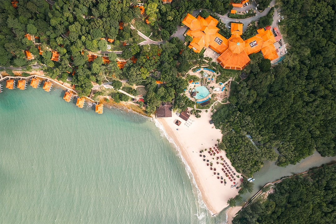 Aerial view of Berjaya Langkawi Resort with overwater chalets along the coastline surrounded by lush rainforest and turquoise sea.