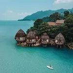 Overwater villas at The Ritz-Carlton Langkawi set above clear turquoise sea with a person paddleboarding nearby and forested hills in the background.