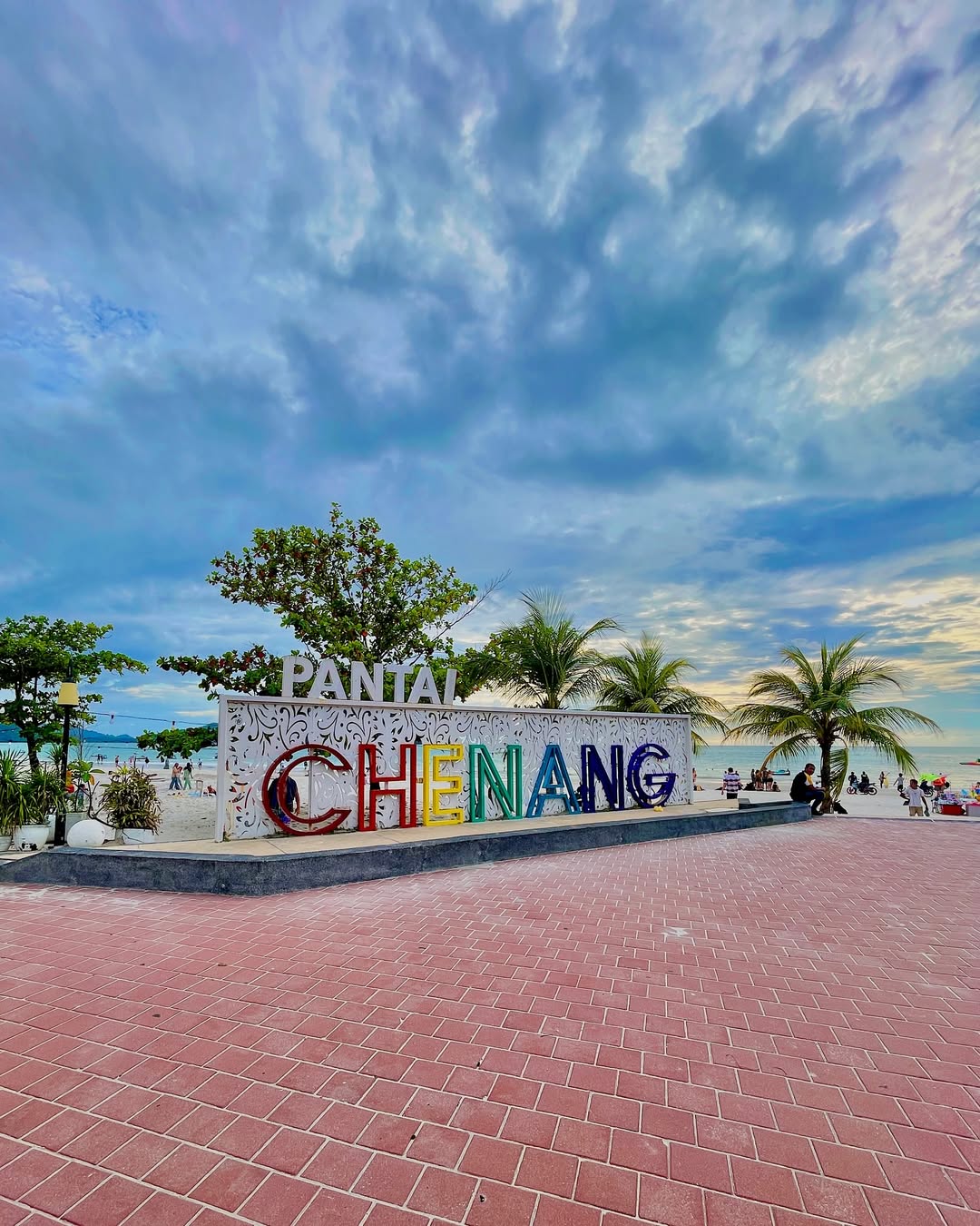 Pantai Cenang sign in Langkawi with colorful lettering, palm trees, and beachgoers by the sea under a cloudy sky.