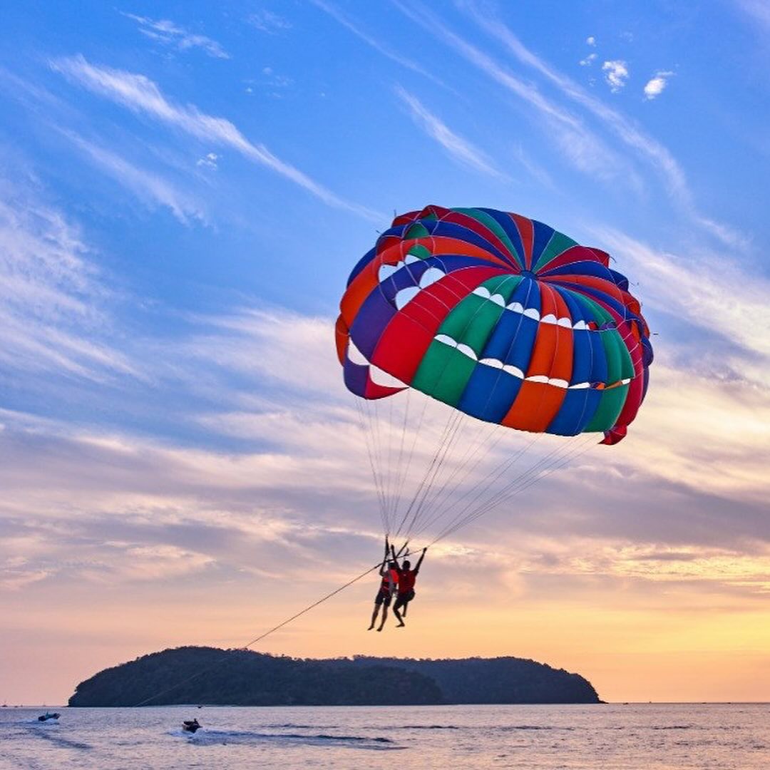 Parasailing activity at Pantai Cenang Langkawi with two people flying above the sea under a colorful parachute during sunset.