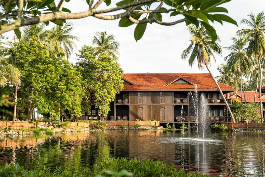 Pelangi Beach Resort & Spa Langkawi featuring traditional Malay-style wooden buildings overlooking a tranquil lagoon, tropical palm trees, lush gardens, and a decorative water fountain in a peaceful resort setting.