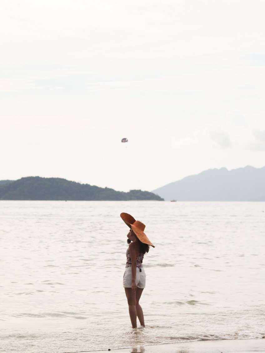 Woman standing in shallow water at Pantai Tengah Langkawi wearing a sun hat, looking out toward the sea and distant islands.