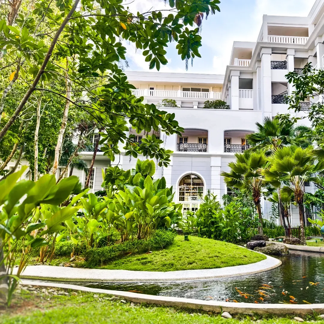 The Danna Langkawi featuring elegant colonial-inspired architecture surrounded by lush tropical gardens, manicured lawns, ornamental ponds with koi fish, and a serene luxury resort atmosphere.