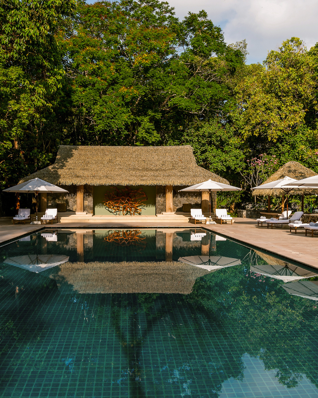The Datai Langkawi featuring a luxurious infinity-style swimming pool surrounded by lush rainforest, elegant sun loungers, tropical landscaping, and a traditional thatched pavilion in a tranquil nature retreat.