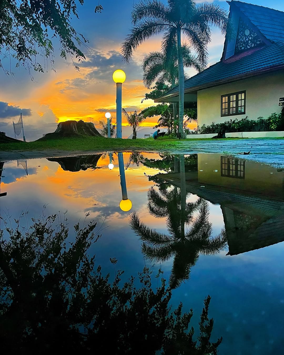 The Frangipani Langkawi Resort & Spa featuring traditional Malay-inspired beachfront villas, tropical palm trees, landscaped gardens, and a stunning sunset reflected in a tranquil water feature along Pantai Tengah.