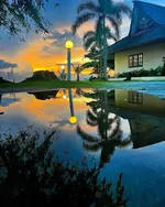 The Frangipani Langkawi Resort & Spa featuring traditional Malay-inspired beachfront villas, tropical palm trees, landscaped gardens, and a stunning sunset reflected in a tranquil water feature along Pantai Tengah.
