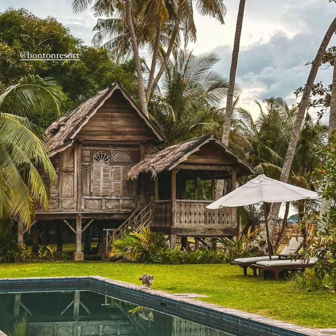 Traditional wooden Malay house on stilts beside a swimming pool at Bon Ton Resort, surrounded by palm trees and tropical greenery in Langkawi.