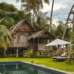 Traditional wooden Malay house on stilts beside a swimming pool at Bon Ton Resort, surrounded by palm trees and tropical greenery in Langkawi.