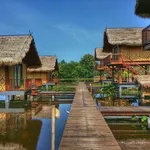 Traditional wooden stilt houses with thatched roofs connected by a wooden walkway over water at The Gemalai Village, surrounded by lush greenery in Langkawi.